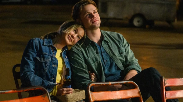 Eine Frau lehnt sich mit dem Kopf auf die Schulter eines Mannes, davor rote Stühle