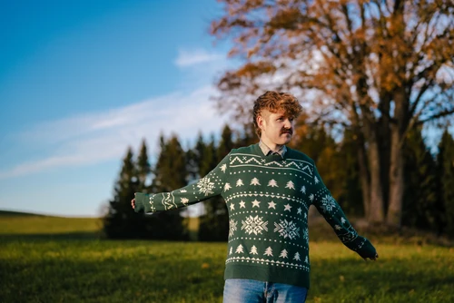 Mann mit Locken und Schnauzer in grünem Weihnachtspullover mit weißem Muster steht in der Natur und streckt die Arme zur Seite