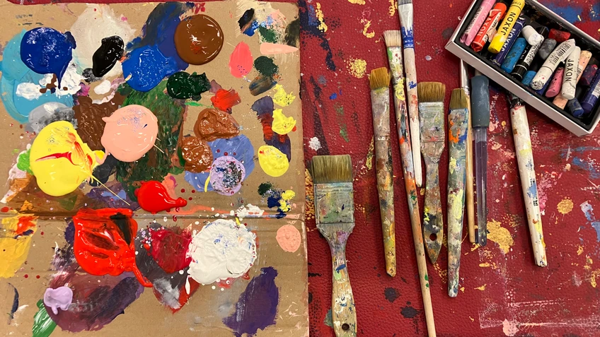 Farbpalette mit bunten Acrylfarben, daneben mehrere farbbespritzte Pinsel und eine Schachtel mit Pastellkreiden auf einer kreativ genutzten Arbeitsfläche.