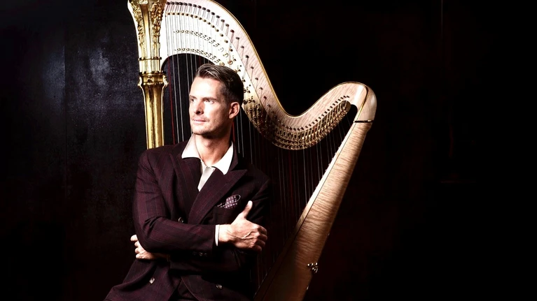 Photo of harpist Xavier de Maistre, elegantly dressed in a dark room against a black background. Directly behind him stands his instrument, the harp, against which he leans slightly.