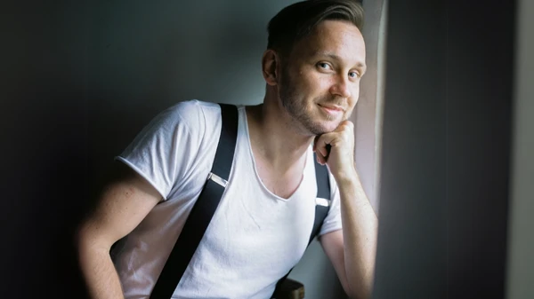 Mann in weißem T-Shirt und Hosenträgern sitzt an einem Fenster und stützt sich auf seine linke Hand
