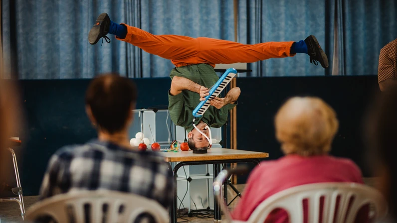 Ein Artist spielt kopfüber im Handstand auf einem Tisch eine Melodica, während vor ihm mehrere ältere Zuschauerinnen und Zuschauer in Plastikstühlen sitzen und zuschauen.