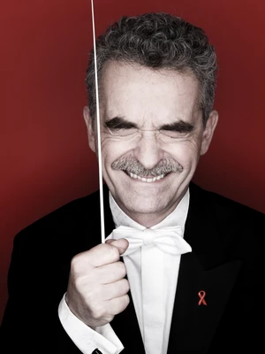 Mann mit grauen Locken und Schnauzbart und weißer Fliege, kneift die Augen zusammen und hält einen Taktstock neben sein Gesicht