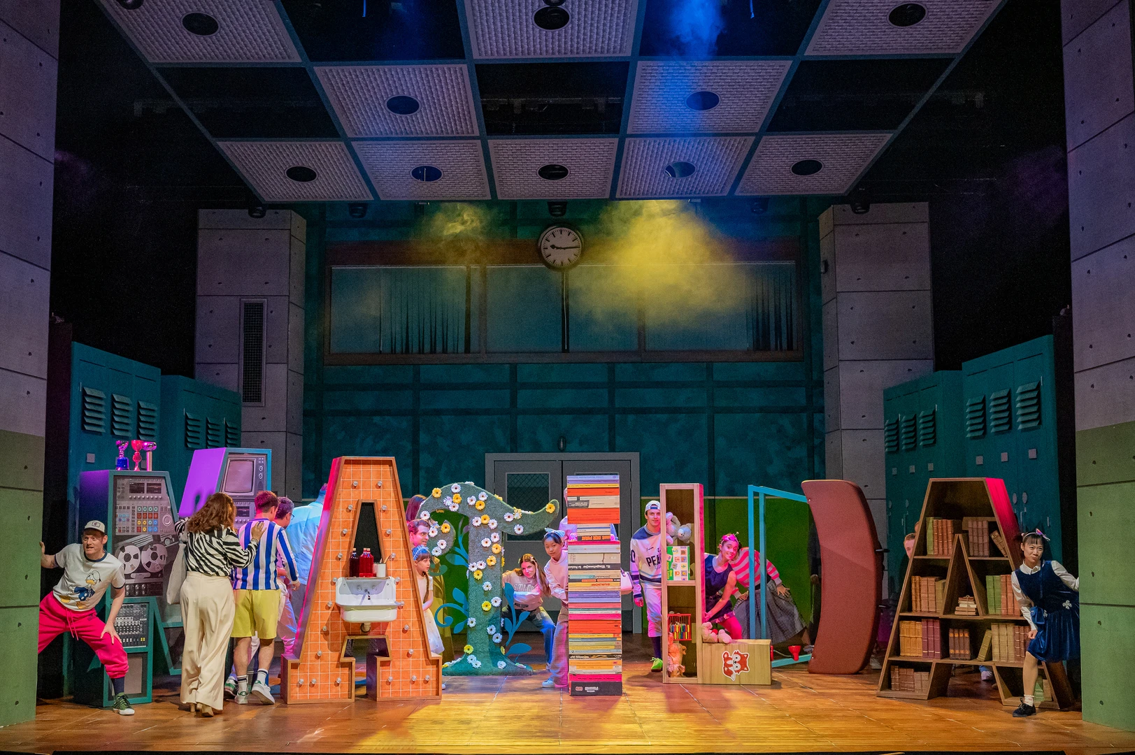 Bühnenbild mit großen bunten Buchstaben, die „MATILDA“ formen. Mehrere Darsteller in farbenfrohen Kostümen stehen und sitzen zwischen den Buchstaben, helle Beleuchtung und Bücherregale im Hintergrund.