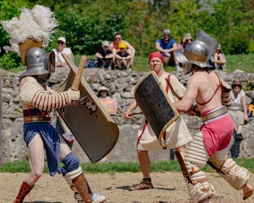 Two gladiators are fighting in the arena on sandy ground. They are wearing helmets, shields, and wooden swords. The referee is in the background. 