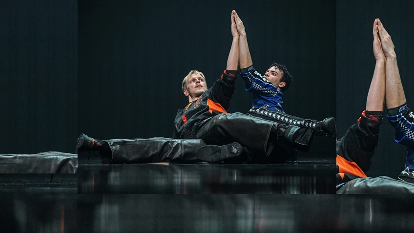 Performancefoto von "Trailer Park", choreografiert von Moritz Ostruschnjak, getanzt von tanz mainz. Die Tänzer:innen performen eine Figur am Boden, ihre Hände miteinander haltend, oversized Sporttrikots tragend.