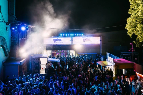 Menschenmenge vor dem Eingang eines nächtlichen Musikfestivals mit Lichtinstallation, Foodtrucks und Bühnenbeleuchtung.