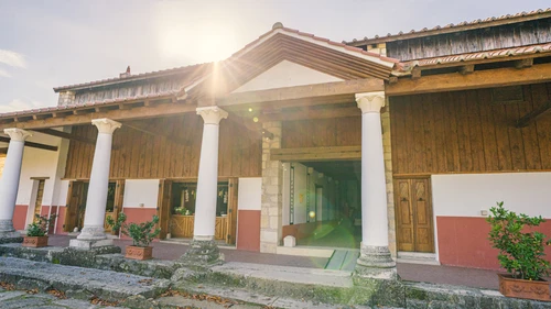 Exterior view of the entrance to the Roman baths. White columns support the canopy. The sun glare over the gable. 