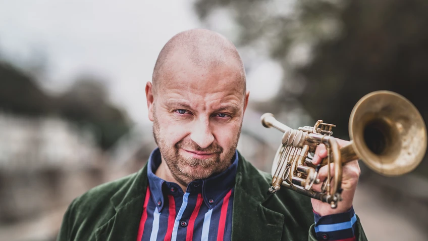 Thomas Gansch mit Stopelglatze, kurzem Bart und farbig gestreiftem Hemd unter einer grünen Samtjacke blickt selbstbewusst in die Kamera, während er eine goldene Trompete locker über der Schulter hält.