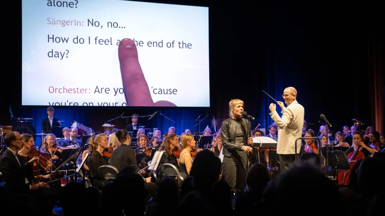 Foto von "Orchesterkaraoke", auf der Bühne ein Orchester, dahinter wird offenbar ein Songtext projiziert, eine Person steht an einem Mikrofon und singt.