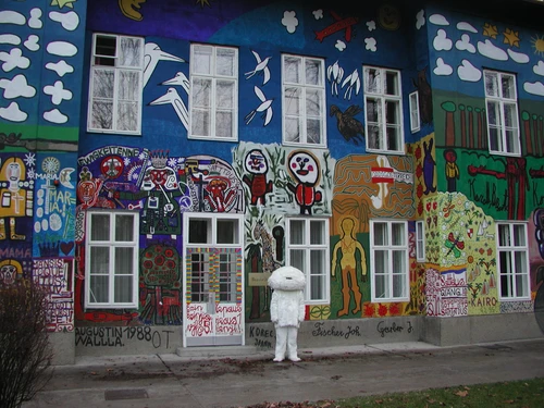 Fassade eines Gebäudes, komplett bedeckt mit farbenfrohen, großflächigen Wandmalereien aus Figuren, Mustern und Symbolen. Vor dem Eingang steht eine Person in einem weißen, flauschigen Kostüm.