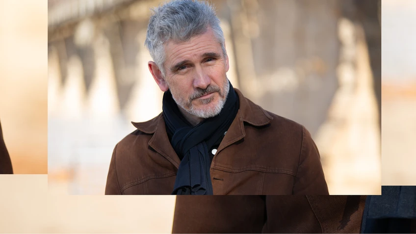 A photo of musician Curtis Stigers wearing a brown jacket and a black scarf, smiling at the camera.