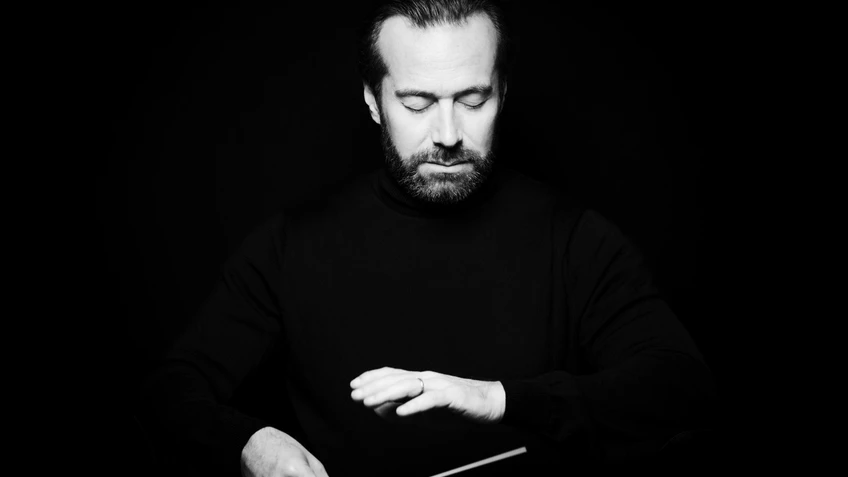 Ein Mann mit Dirigierstock vor schwarzem Hintergrund, er hat die Augen geschlossen