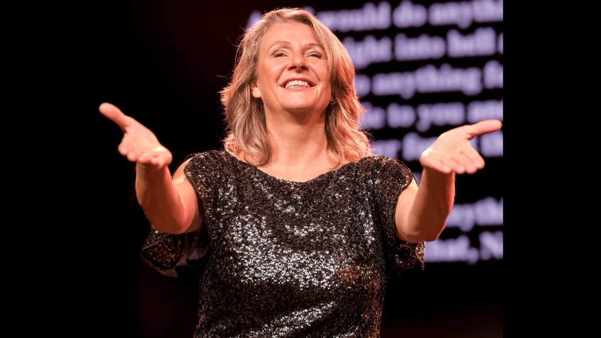 Eine Frau in schwarz glitzerndem Kleid lächelt ins Publikum und streckt die Arme einladend aus