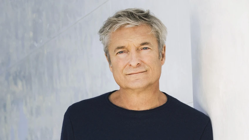 Mann in dunklem Pullover mit grauen Haaren an einer weißen Wand