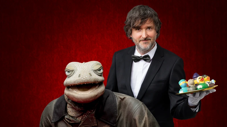 Michael Hatzius steht im Smoking vor einem roten Hintergrund und hält ein Tablett mit bunten Plastikenten, während neben ihm die Echse in brauner Kleidung sitzt und eine Zigarre in der Hand hält.