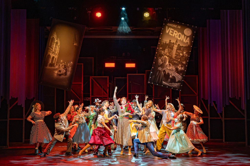 A large ensemble scene on a theatre stage featuring numerous performers gathered in a tight group at the centre of the stage. Many of the performers have their arms stretched upwards or are striking dynamic poses. The stage is illuminated by warm red and orange lighting, as well as occasional blue highlights. In the background, two large banners or posters hang diagonally above the scene, whilst tall curtains and other stage elements frame the space. The patterned stage floor enhances the effect of the central choreography. The composition draws the eye to the group’s collective movement and conveys the impression of a festive or concluding ensemble performance.
