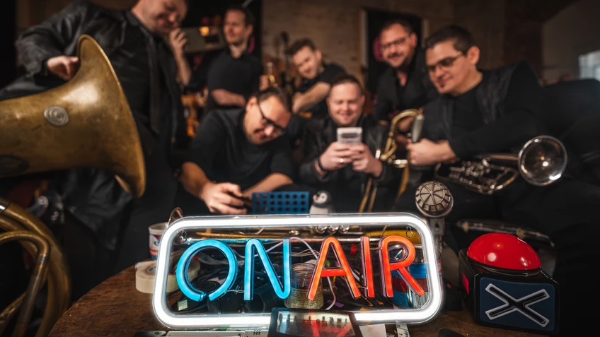 Musiker sitzen rund um ein Leuchtschild mit der Aufschrift "on air", Instrumente und andere Gegenstände unscharf