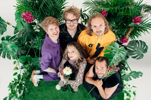 Vier Männer und eine Frau in kreativer Kleidung in einer Kulisse aus verschiedenen Pflanzen, weißer Hintergrund