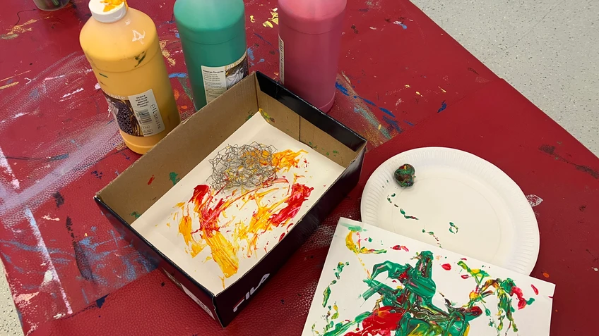 Basteltisch mit offenen Farbtuben in Gelb, Grün und Rot, einer Kartonschachtel mit bemaltem Papier und einer weiteren abstrakten Farbarbeit auf einem Teller.
