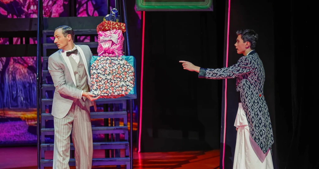 links steht ein Mann mit grauem Anzug hat einen Geschenkestapel in den Händen, gegenüber auf der echten Seite zeigt ein junger Mann auf die Geschenke