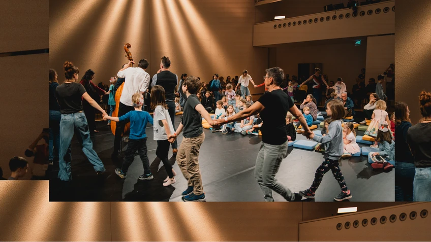 Foto von Big Bang - dem Musikabenteuer für junges Publikum. Zu sehen sind Kinder und Erwachsene, die Ringelreihen tanzen, begleitet von Musiker:innen, die Live-Musik spielen. 