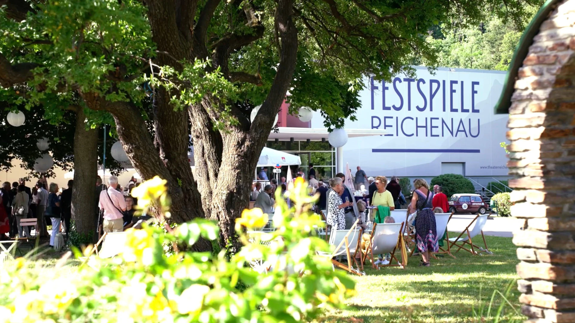 Besucherinnen und Besucher bei sonnigem Wetter im Gartenbereich der Festspiele Reichenau vor dem Festspielgebäude.