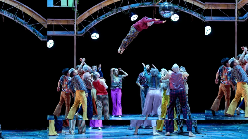 A still from the performance *Beauséjour* by Mourad Merzouki, performed by Compagnie Käfig. The photograph shows the dancers and acrobats on stage in colourful costumes; one is being thrown through the air, whilst other dancers stand ready to catch them.