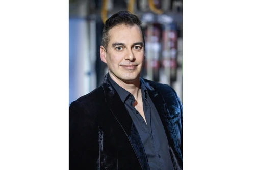 Eine Person trägt ein dunkles Samt-Sakko über einem dunklen Hemd; der Hintergrund zeigt verschwommene Studioelemente. Das Gesicht ist absichtlich unscharf und nicht erkennbar.