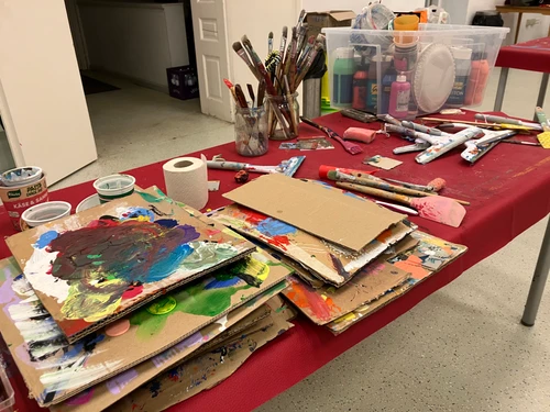 Kreativer Arbeitstisch mit bemalten Kartonplatten, Pinseln, Farben und Malutensilien in einem Atelier- oder Workshopraum.