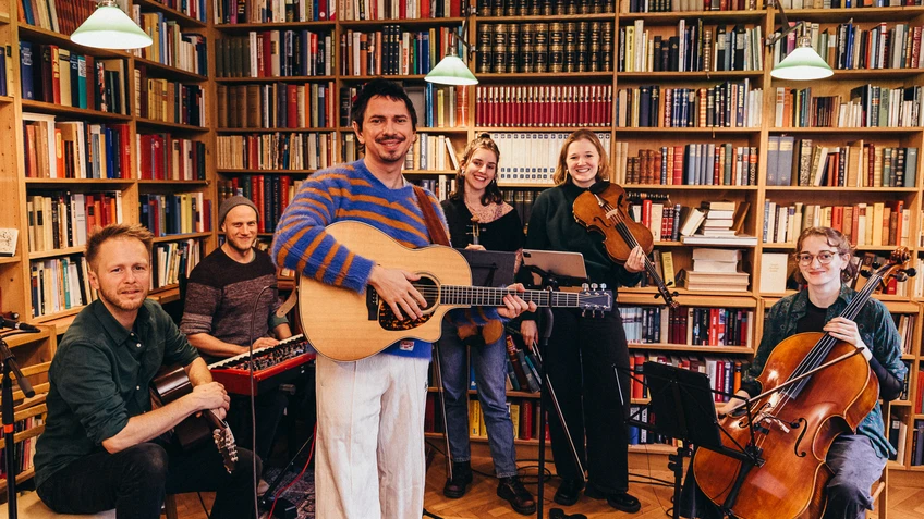 Ein Gitarrist in blau-orange-gestreiftem Pullover in einer kleinen Bibliothek mit 5 MusikerInnen und ihren Instrumenten