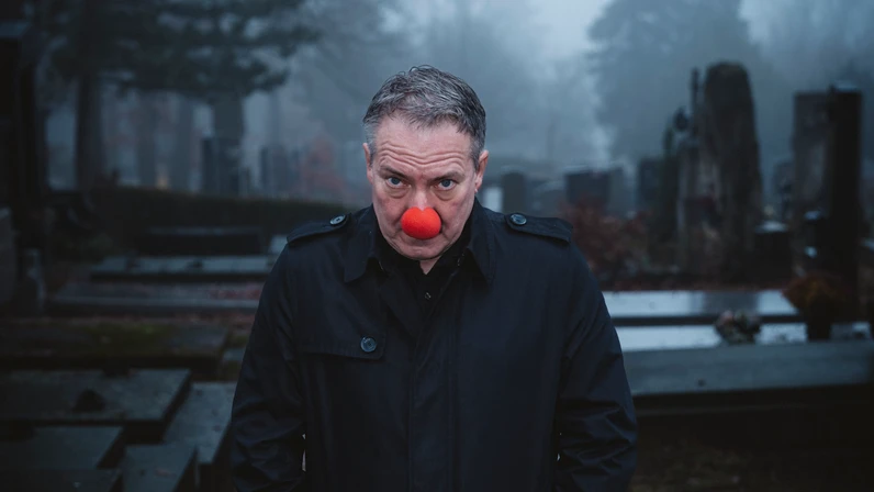 Robert Palfrader steht mit schwarzem Mantel und roter Clownsnase nach vorn gebeugt und ernst blickend auf einem nebligen Friedhofsweg zwischen Gräbern.