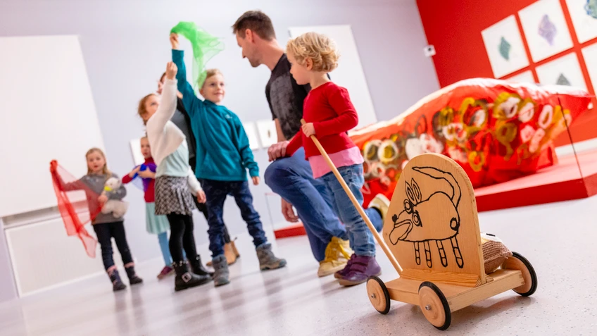 Kinder und Erwachsene bewegen sich gemeinsam durch einen Museumsraum mit spielerischen und kreativen Objekten.