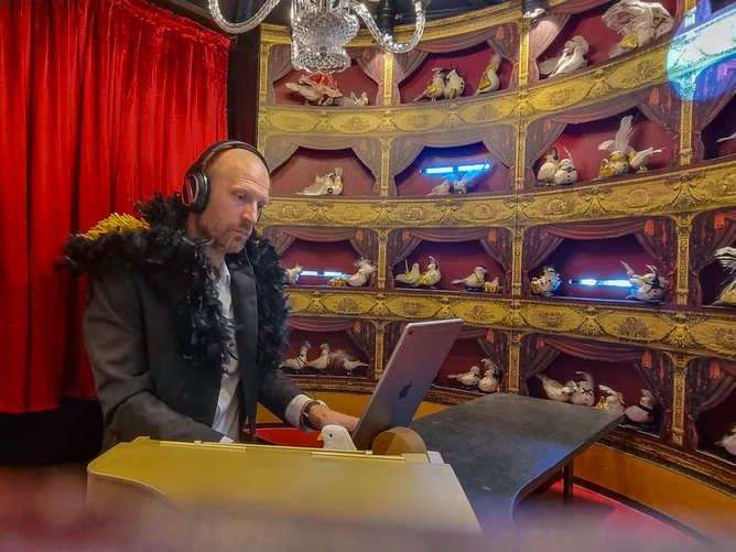 The project "Duvekot" – the Danish word for "dovecote" – can be seen in an interior view; inside "the smallest concert hall in the world" with theatre-boxes in which handmade birds sit, and in the middle a man with headphones who is apparently listening to music.