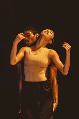 Performancefoto der Choreografie "SWAY" von Medhi Walerski. Zu sehen sind  zwei Tänzer:innen des Ballet BC, gekleidet in schwarzer Hose und weißem Shirt, vor schwarzem Bühnenhintergrund, in einem körperlich-engen Tanzschritt, beleuchtet mit gelbem Bühnenlicht. 