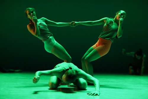 Performancefoto von "Sadeh21", choreografiert von Ohad Naharin. Zu sehen sind Tänzer:innen der Batsheva Dance Company in bunten Kostümen in Gruppenchoreografien tanzend auf einer grün beleuchteten Bühne.