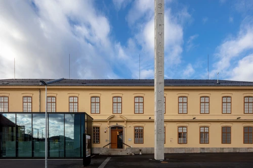 Eingang der Kunsthalle Krems im historischen gelben Fabriksgebäude; Glasfoyer und hoher Betonpylon mit Schriftzug KUNSTHALLE KREMS davor.
