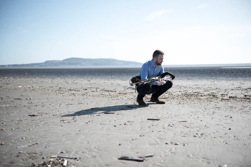 Foto von Musiker Mark Redmond mit seinem Instrument, mit Uilleann Pipes, zu sehen am Strand, hinter ihm das Meer.