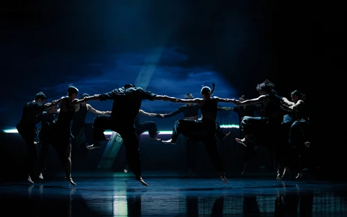 Szenenfoto von "All Ears", einer Performance vom Cloud Gate Dance Theatre of Taiwan, choreografiert von Cheng Tsung-lung. Zu sehen sind die schwarz gekleideten Tänzer:innen, die im Kreis angeordnet sind, sich an den Händen haltend und springend.