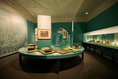 Blauer Raum mit blauem Tisch, darauf sind mehrere Glasvitrinen mit verschiedenen Büchern und Dokumenten. Auf der rechten Wand eine lange Wandvitrine mit verschiedenen Gefäßen.