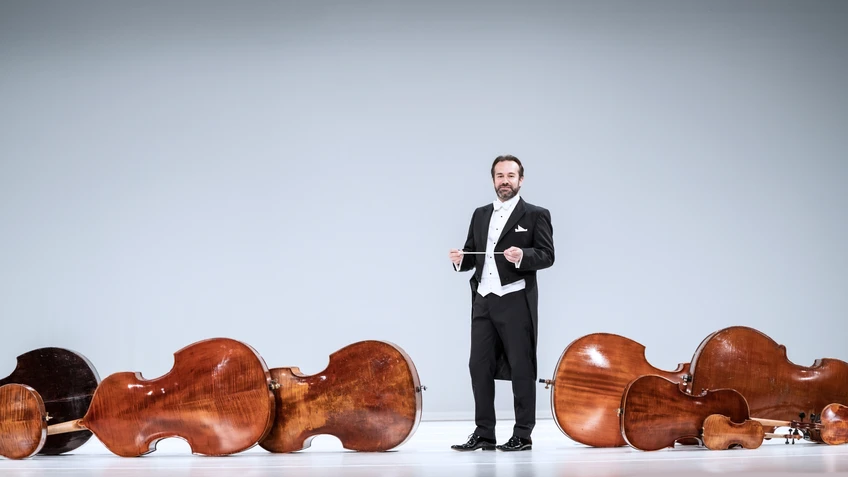 Fabien Gabel in a tailcoat in front of a white background, surrounded by orchestral instruments on the floor. 