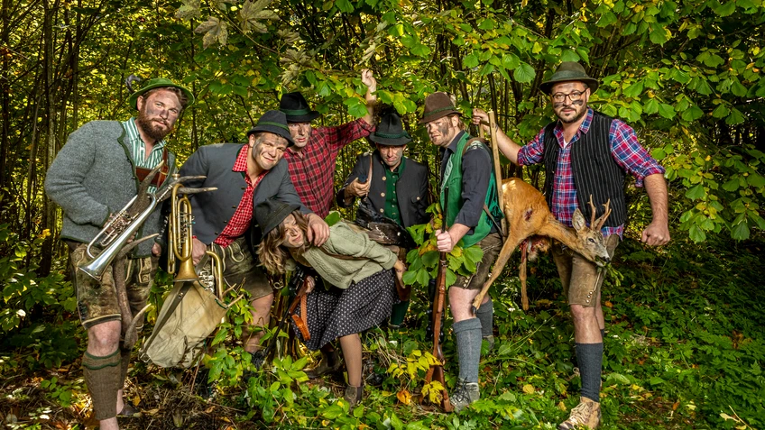 Musikgruppe in Tracht mit Tarnfarbe im Gesicht mit Instrumenten in grünem Gebüsch