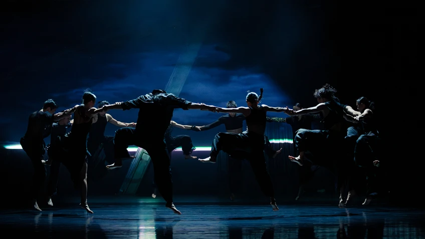 Szenenfoto von "All Ears", einer Performance vom Cloud Gate Dance Theatre of Taiwan, choreografiert von Cheng Tsung-lung. Zu sehen sind die schwarz gekleideten Tänzer:innen, die im Kreis angeordnet sind, sich an den Händen haltend und springend.