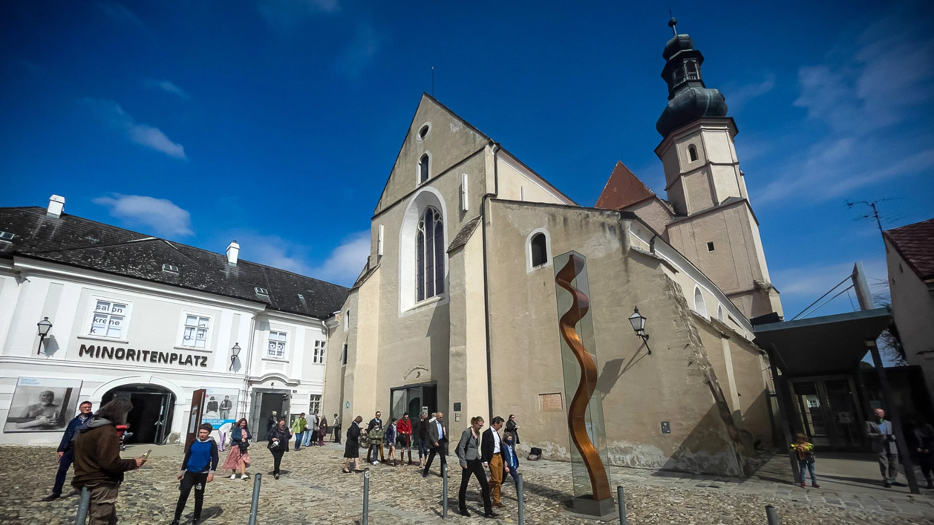 Historischer Platz mit gotischer Kirche, moderner Skulptur und Fußgängern bei sonnigem Wetter.