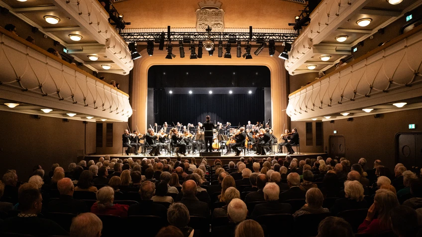 Saal eines Theaters voll mit Publikum, auf der Bühne ein Symphonieorchester im Scheinwerferlicht