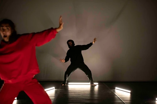Performancefoto von "Rotkäppchen", choreografiert von Sylvain Huc. Zu sehen sind zwei Tänzer:innen, eine:r davon in einem knallroten Sportanzug, der:die andere in einem schwarzen Sportanzug.