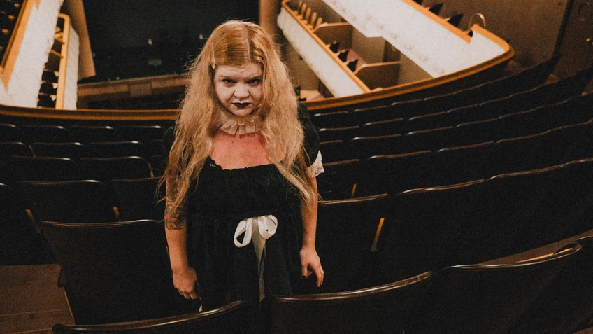 Frau blass geschminkt mit blonden Haaren und dunklen Lippen im Rang eines Theaters