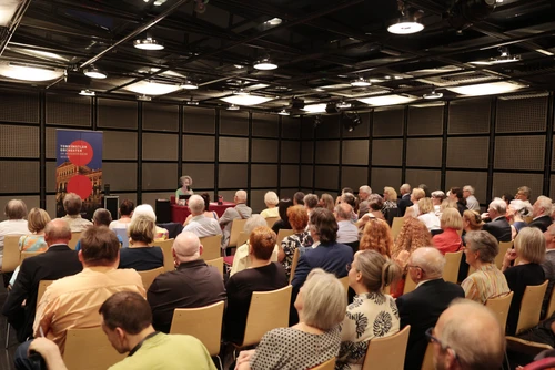 Dunkler Sall, in dem eine Frau an einem Tisch sitzt und vor Publikum vorträgt