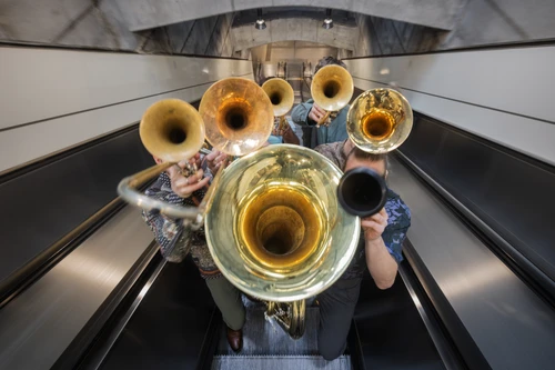 Gruppenfoto vom Bläserensemble "Federspiel", aufgenommen auf einer Rolltreppe in einer U-Bahnstation. Die Musiker stehen dicht beisammen und halten ihre Instrumente vor ihre Gesichter, sodass diese verdeckt bleiben.