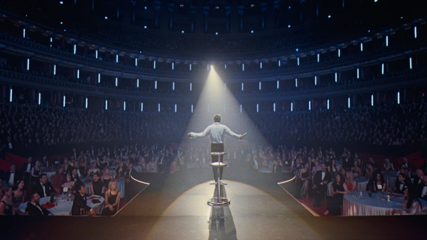 Eine Person von Hinten auf einer Bühne, dahinter ein großer Saal schwach beleuchtet voller Publikum